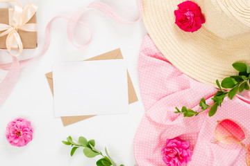 Flat lay fashion office table desk. Female workspace with blank paper card, craft envelope, straw hat, rose flower buds, gift box, branches on white background. Top view feminine background