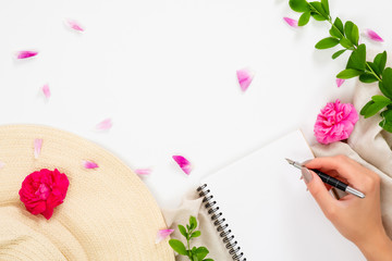 Flat lay, top view office table desk. Workspace with straw hat, blank paper notepad, human hand holding pen, pink rose flower buds and petal on white background.