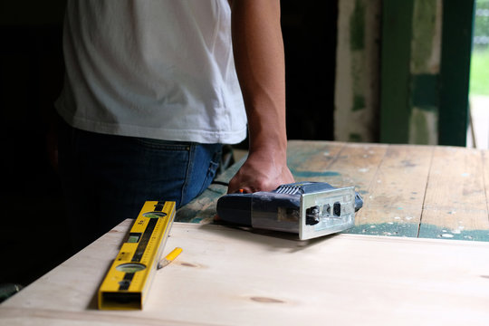 Hands of carpenter holding electric jigsaw lying on workbench with spirit level and yellow pen. DIY workwood concept.