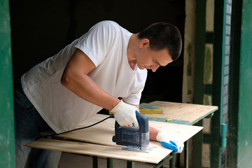 Young carpenter is sawing a plywood sheet with electric jig saw machine in carpentry workshop. Workwood DIY concept.