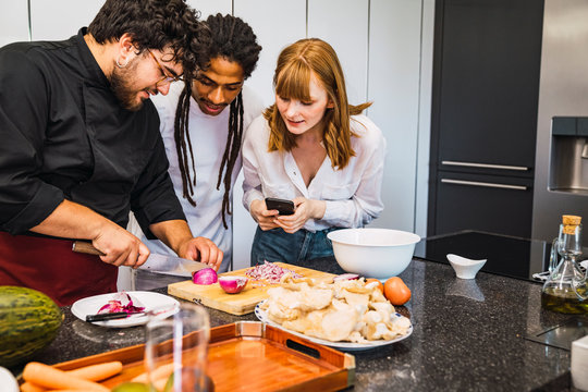 Professional cook teaching a black guy and a red Caucasian girl how to cook. She carries her mobile in her hand to make a video. Modern kitchen.