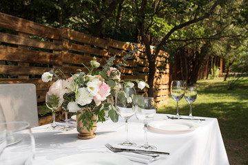 Wedding table setting decorated with fresh flowers in a brass vase. Wedding floristry. Banquet table for guests outdoors with a view of green nature. Bouquet with roses, eustoma and eucalyptus leaves