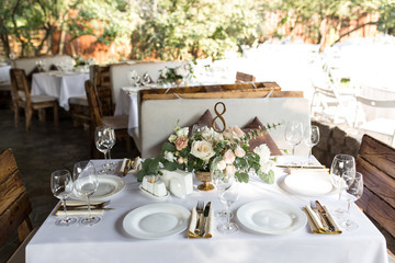Wedding table setting decorated with fresh flowers in a brass vase. Wedding floristry. Banquet table for guests outdoors with a view of green nature. Bouquet with roses, eustoma and eucalyptus leaves