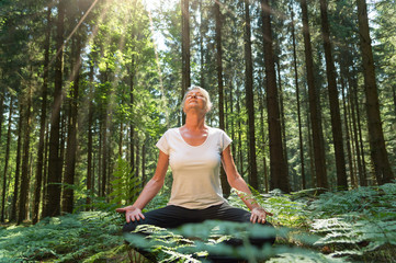 Experience the forest while bathing in the forest (Shinrin Yoku) with all her senses. A 50 year old blonde woman sits cross legged relaxed. She fuels the sun and the atmosphere of the forest.