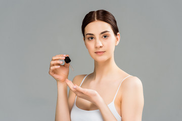 attractive brunette woman holding pipette and looking at camera isolated on grey