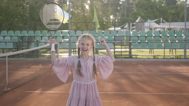 Cute little smiling happy girl with a tennis racket in her hands standing on the tennis court looking into the camera. Grandfather and grandmother rejoice at their granddaughter showing thumb up.