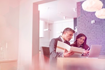 Mature male realtor explaining document with laptop to female buyer at table in apartment