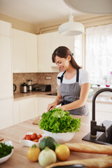 Beautiful smiling dedicated Caucasian brunette in apron standing in kitchen and chopping mushrooms. On table are lots of vegetables. Cooking at home concept.