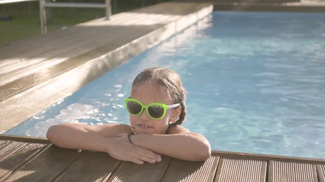 Portrait pretty funny girl in yellow sunglasses looking in the camera smiling, looking out of the pool, holding on to the edge. Child having fun in the hotel complex. Recreation and leisure outdoors.