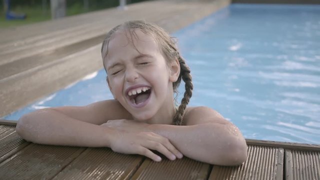 Portrait cute funny girl with pigtails laughing looking in the camera. The child showing thumb up looking out of the pool, holding on to the edge. Preteen kid having fun in the hotel complex