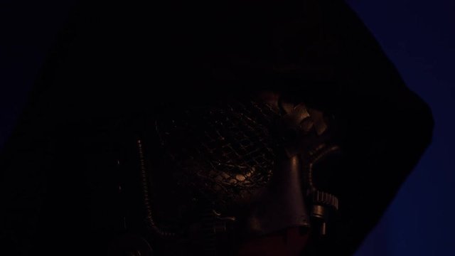 Close-up on the face of a man in a black hood and a leather mask lit by light. The mask has a grid, gears and patches in the form of puzzle pieces. The dark room, beam slides over mask. Puzzle concept