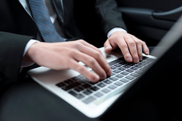 View of hands of businessman in a suit and tie, who is sitting in a car, with laptop on knees and typing on keyboard.