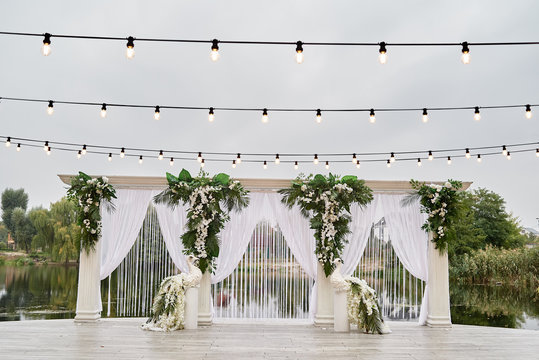 Luxury wedding arch decorated with palm leaves, orchid flowers, floral peacocks and bulbs garland outdoors, copy space. Wedding ceremony near water in park
