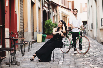 Fototapeta na wymiar A loving couple in the old city. A woman in a black dress sits on a chair. The man behind her stands with a green bike