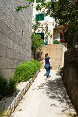 Fototapeta na wymiar Young girl walking through a narrow alley in a residential area in the mediterranean towns. Flowers in the street in summer resort. Red head with pony tail