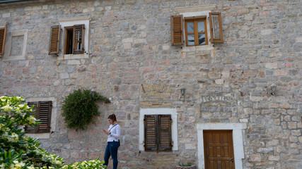 Fototapeta na wymiar Girl with white long shirt sending a message in smart phone and stone house with flowers in the facade. Woman typing in cell phone and wood windows in mediterranean town