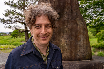 A middle aged man travels in Kyoto, Japan, to the beautiful Arashiyama area, and poses in front of an old stone monument.