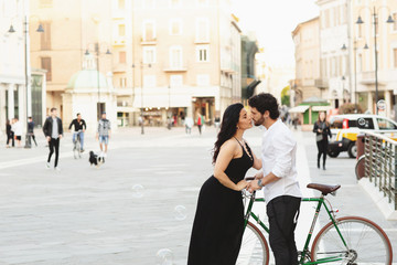 Man and woman with a bicycle are kissing in the old city. Around them are water bubbles. Love story in Rimini, Italy