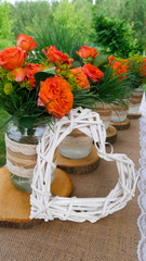 Retro decoration of the table with orange roses bouquets in glass jars and wattle white heart in the garden