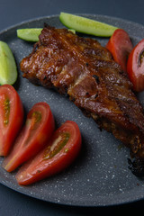 baked pork ribs with honey lie with fresh sliced ​​tomatoes and cucumbers on a dark gray concrete tabletop