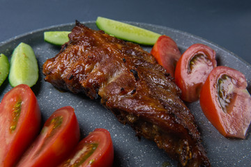 baked pork ribs with honey lie with fresh sliced ​​tomatoes and cucumbers on a dark gray concrete tabletop