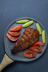 baked pork ribs with honey lie with fresh sliced ​​tomatoes and cucumbers on a dark gray concrete tabletop