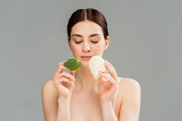 young brunette woman with acne on face holding broccoli and cauliflower isolated on grey