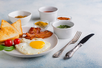 Fried egg with bacon, toast and tomatoes decorated with a leaf of basil on a white background with a knife and fork. English breakfast. with copy space for your text