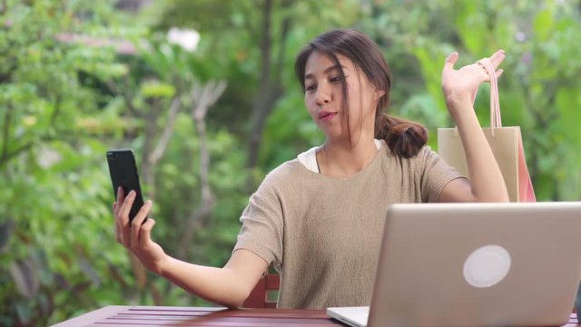 Asian woman using mobile phone selfie post in social media, female relax feeling happy showing shopping bags sitting on table in the garden in morning. Lifestyle women relax at home concept.