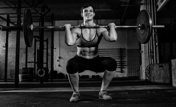 Schöne muskulöse Frau mit kurzen Haaren beim Workout mit der Langhantel. Die junge athletische Sportlerin trainiert Kniebeugen mit der Langhantel. Krafttraining mit Gewichten. Schwarz und weiß.