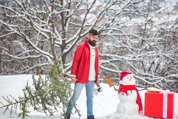 Bearded man with snowman is carrying Christmas tree in the wood. A handsome young man with snow man carries a Christmas tree. Merry Christmas and happy new year greeting card. Snowman with gift.