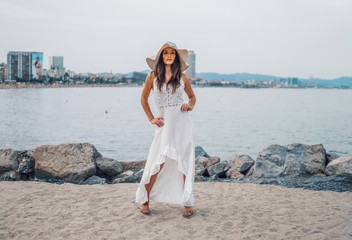 Pretty woman wearing white dress and pamela hat standing over the city beach, sea. Happy summer...