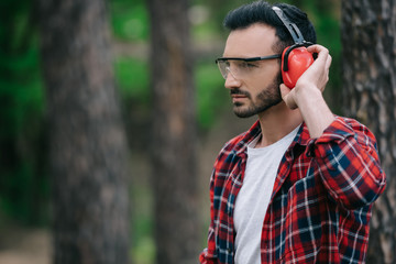 pensive lumberjack touching noise-canceling headphones and looking away in forest