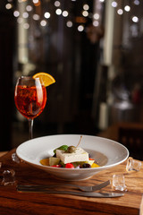 Refreshing summer cocktail Aperol Spritz and greek salad with feta cheese on black background with blurred bokeh lights