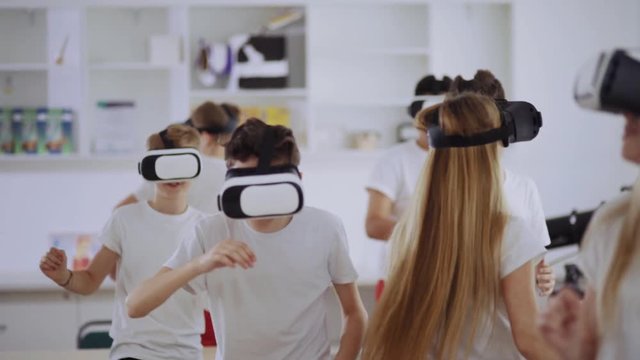 Joyful Schoolchidren Discovering Virtual Reality Wearing Headsets Dancing And Moving In The Middle Of The Classroom During A Lesson.