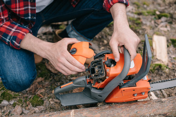 cropped view of lumberer in plaid shirt fixing chainsaw in forest