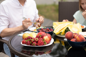 Cropped stock photo of unrecognizable family having picnic with summer fruits at the table outdoors. Various fruit on the table. Peaches, strawberries, cherries, watermelon, apples.