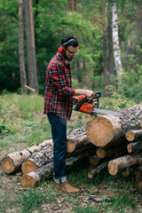 bearded lumberjack in noise-canceling headphones cutting wood with chainsaw in forest