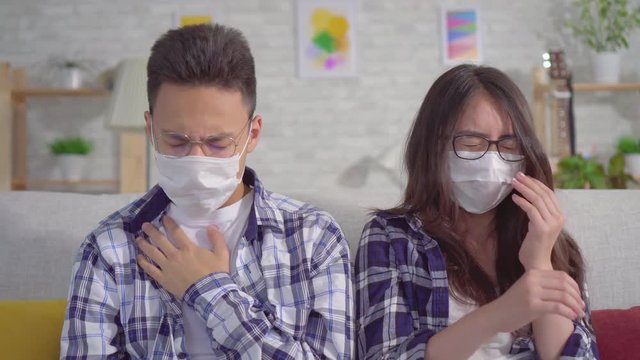Sick of a cold young sian couple sitting in protective medical masks on the couch