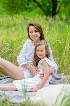 Beautiful young mother and her little daughter in white dress having fun in a picnic. They are sitting on a plaid, hugging and smiling. Maternal care and love. Verticalal photo