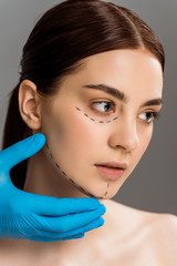 cropped view of plastic surgeon in latex glove touching face of pretty naked woman isolated on grey