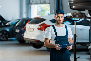 happy bearded car mechanic looking at camera and holding clipboard near cars