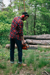 adult bearded man in checkered shirt and earmuffs holding chainsaw in forest