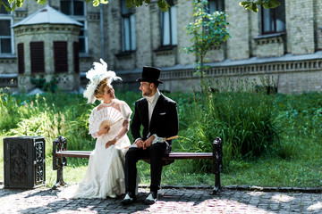 Obraz premium cheerful victorian woman holding fan while sitting with young man on bench