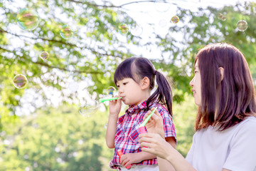 新緑の樹木を背景にシャボン玉で遊ぶ幼い女の子と母親。幸せ、愛情、親子イメージ
