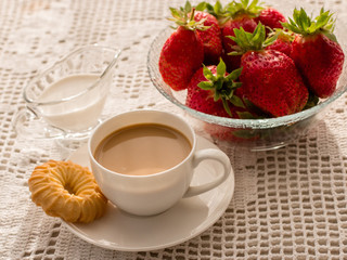 Still life. Coffee with cream in a white cup, round biscuits, a glass plate with red, ripe strawberries on a table covered with a handmade lace tablecloth. Close-up.