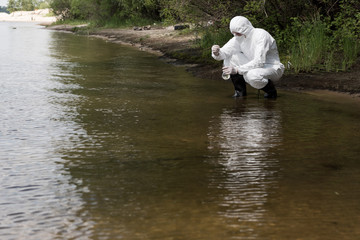water inspector in protective costume, latex gloves and respirator taking water sample at river
