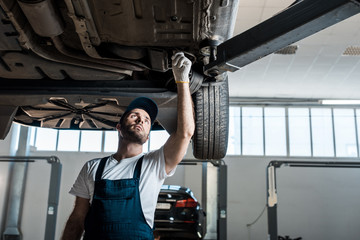 bearded car mechanic in cap looking at automobile in car service