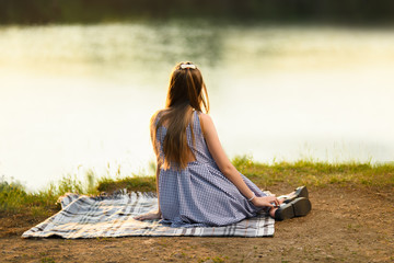 Girl blonde in a light dress sitting on a blanket and looking into the distance at sunset. Girl at sunset. Horizontal photography