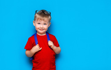 Happy smiling boy in red t-shirt with glasses on his head is going to school for the first time. Child with school bag. Kid on blue background background. Back to school